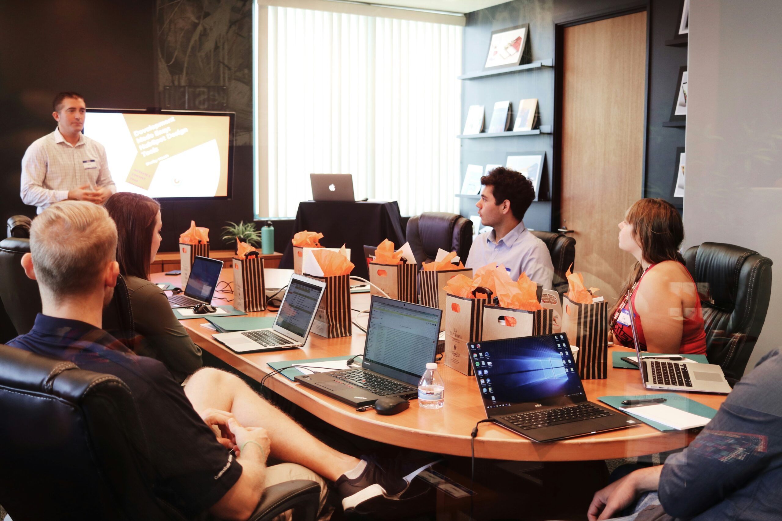A presenter speaking to colleagues seated around a conference table with laptops.