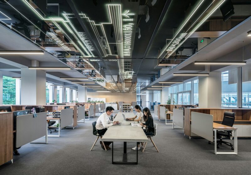 Three people working at laptops around a shared table in a modern open-plan office.