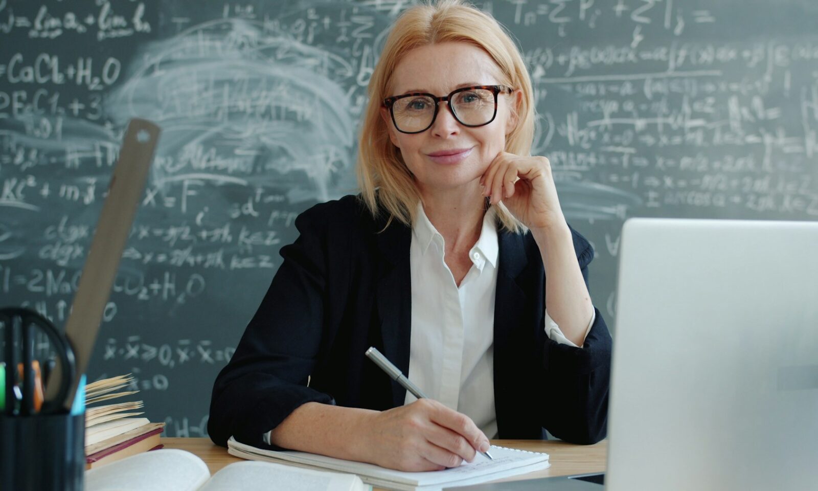 Teacher smiling at a classroom desk with a laptop and books