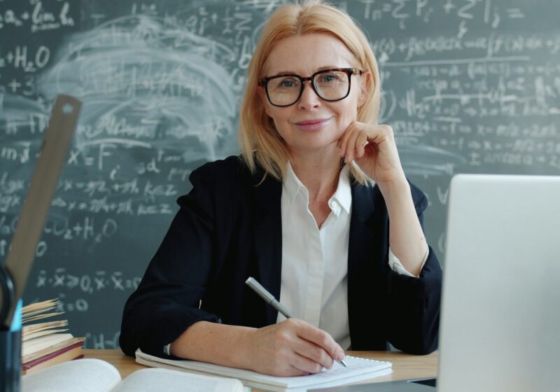 Teacher smiling at a classroom desk with a laptop and books