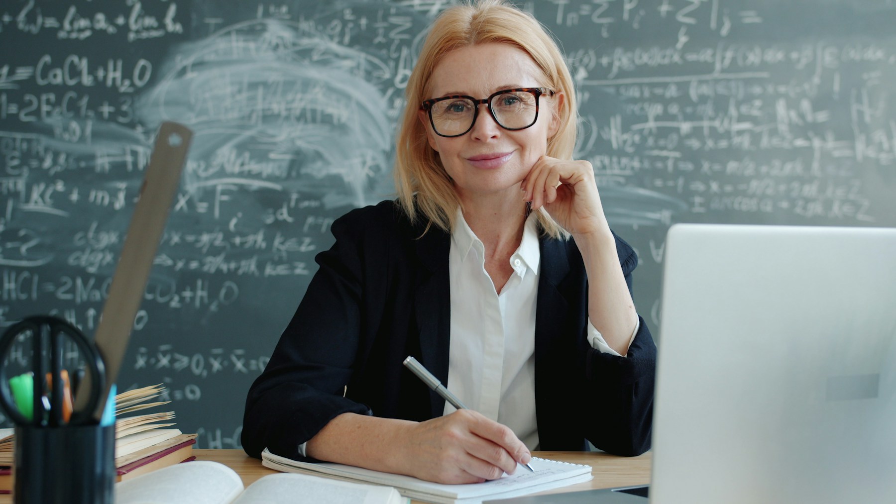 Teacher smiling at a classroom desk with a laptop and books